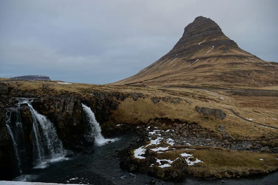 Eine beeindruckende Aussicht auf den Kirkjufell, einen markanten Berg in Island, zusammen mit einem Wasserfall im Vordergrund und grauem Himmel.