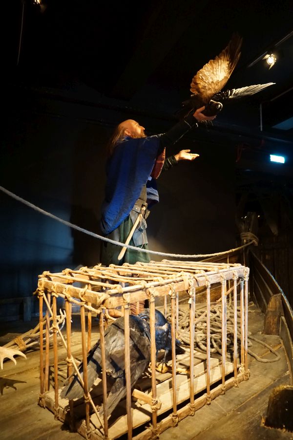 Ein Wikinger in traditioneller Kleidung steht mit ausgebreiteten Armen und hält einen Adler, während ein Rabe in einem Käfig neben ihm sitzt.