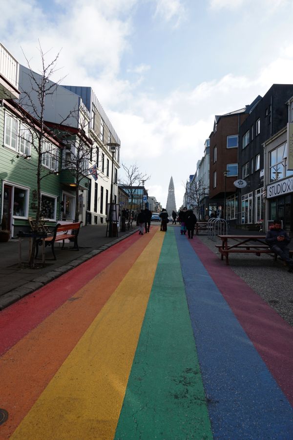 Farbenfrohe Fußgängerzone in Reykjavik mit einem regenbogenfarbenen Streifen auf der Straße und Blick auf die Hallgrímskirkja im Hintergrund.
