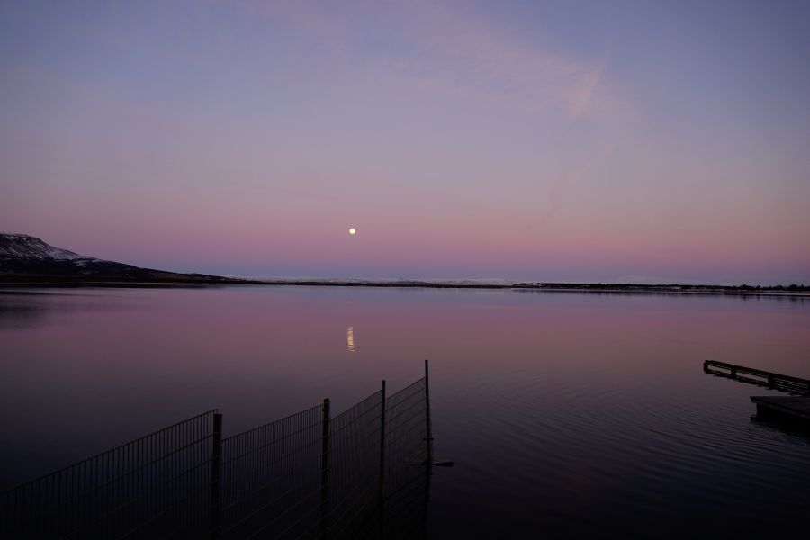 Ein traumhaftes, ruhiges Abendbild eines Sees mit sanften pinken und lila Farbnuancen im Himmel, während der Mond über dem Horizont steht.