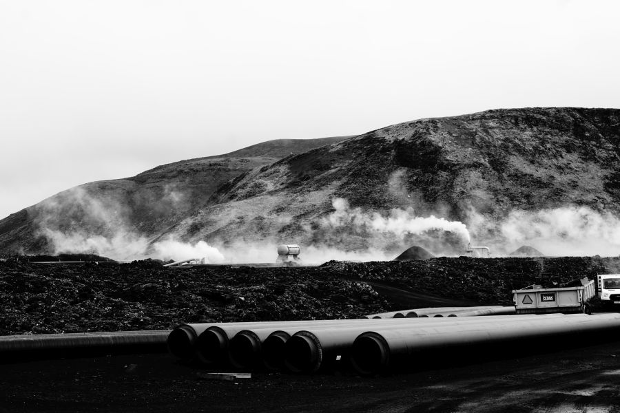 Ein schwarz-weißes Bild von einer geothermischen Landschaft mit dampfenden Hügeln, großen Rohrleitungen im Vordergrund und einer Baustelle in der Nähe.