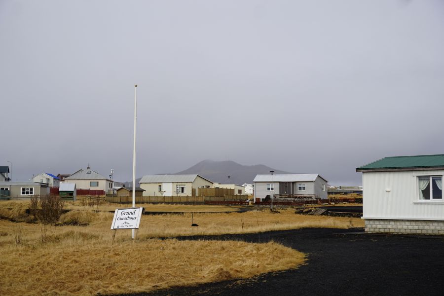 Ein ruhiger Blick auf ein kleines Dorf in Island, mit mehreren Häusern im Vordergrund und einem Hügel im Hintergrund, umgeben von grauem Himmel.