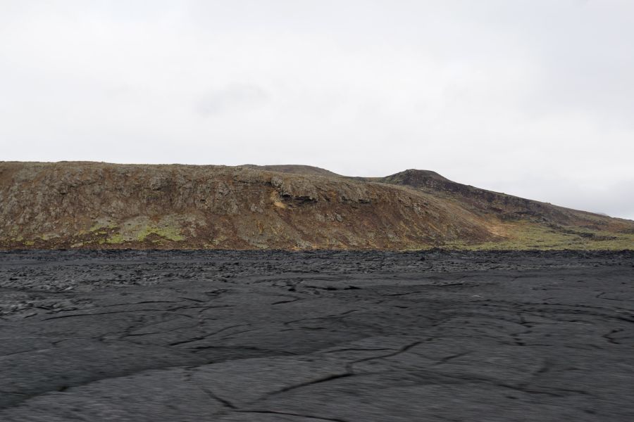 Schwarze Lavafelder mit erdigen Hügeln im Hintergrund unter einem bewölkten Himmel.