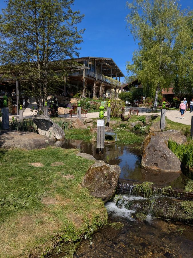 Ein ruhiger Gartenbereich mit einem kleinen Teich, Steinen und Bänken. Im Hintergrund ein Holzgebäude mit viel Glas und von Bäumen umgeben unter blauem Himmel.