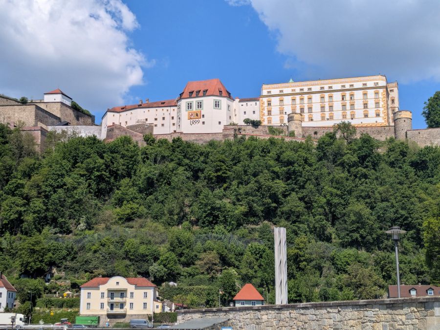 Ein historisches Schloss, das auf einem Hügel thront, umgeben von üppigem Grün und Bäumen, mit blauen Himmel und einigen Wolken im Hintergrund.