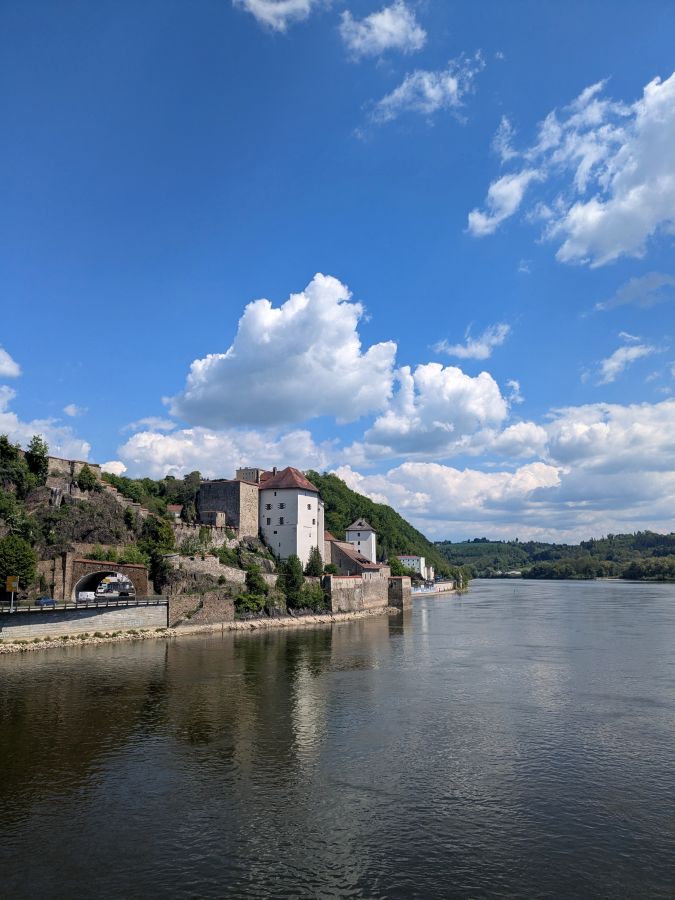 Blick auf eine historische Stadtmauer und Gebäude entlang eines Flusses an einem sonnigen Tag mit Blütenwolken am Himmel.