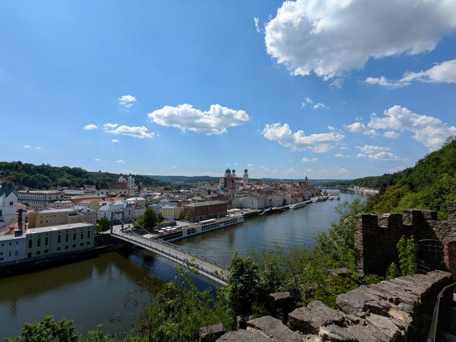 Blick auf die Stadt Passau mit der Donau, umgeben von grünen Hügeln und historischem Stadtbild unter blauem Himmel mit Wolken.