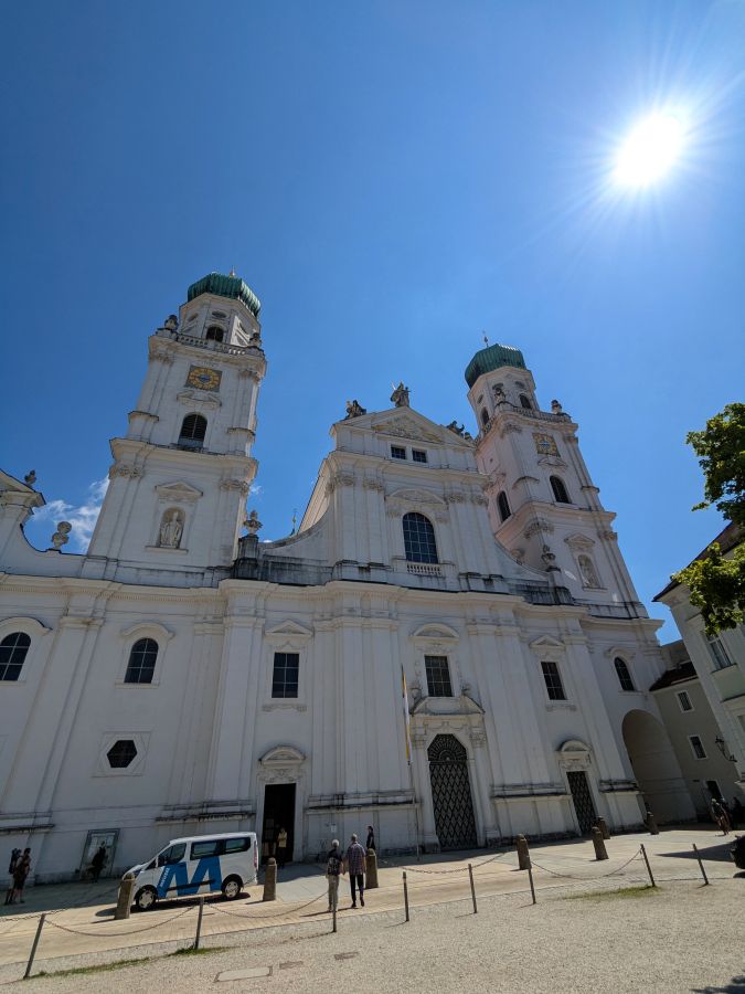 Ansicht der beeindruckenden Stadtpfarrkirche St. Paul in Passau unter einem strahlend blauen Himmel, mit der Sonne im Hintergrund und Menschen, die in der Nähe stehen.