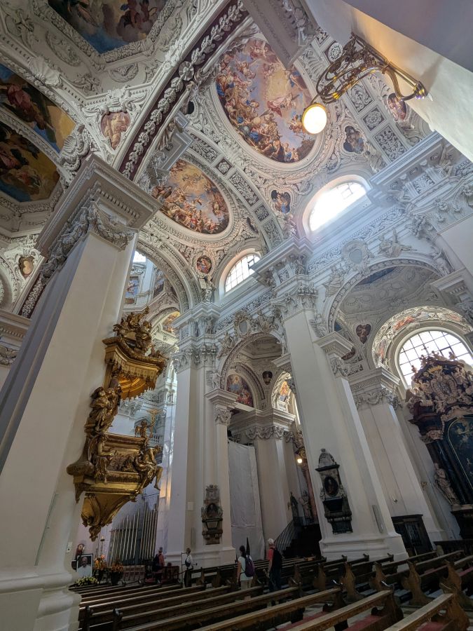 Blick in die kunstvoll gestaltete Decke einer Kirche mit reich verzierten Wänden und bunten Fenstern.