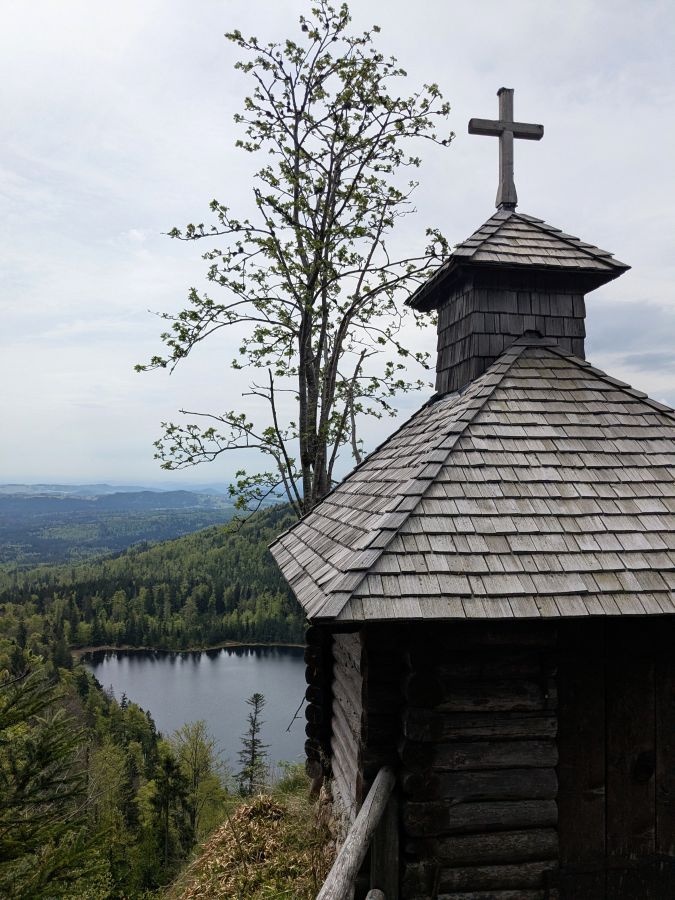 Eine kleine Holzkirche mit Satteldach und Kreuz auf dem Dach, umgeben von Bäumen und einer Aussicht auf einen See und die hügelige Landschaft im Hintergrund.