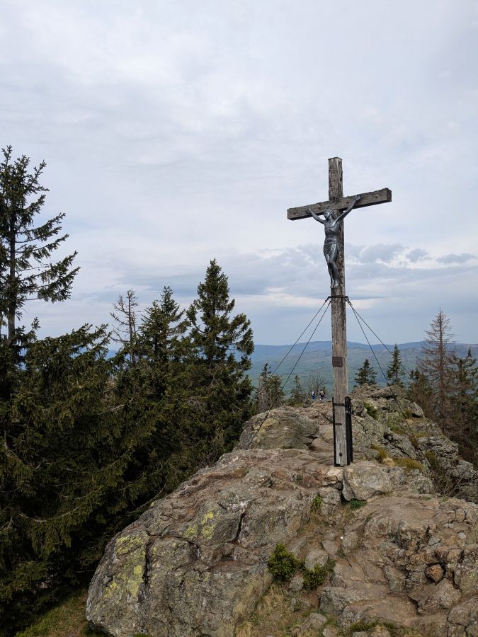 Holzkreuz auf einem Gipfel, umgeben von Kiefern und Aussicht auf die Landschaft.
