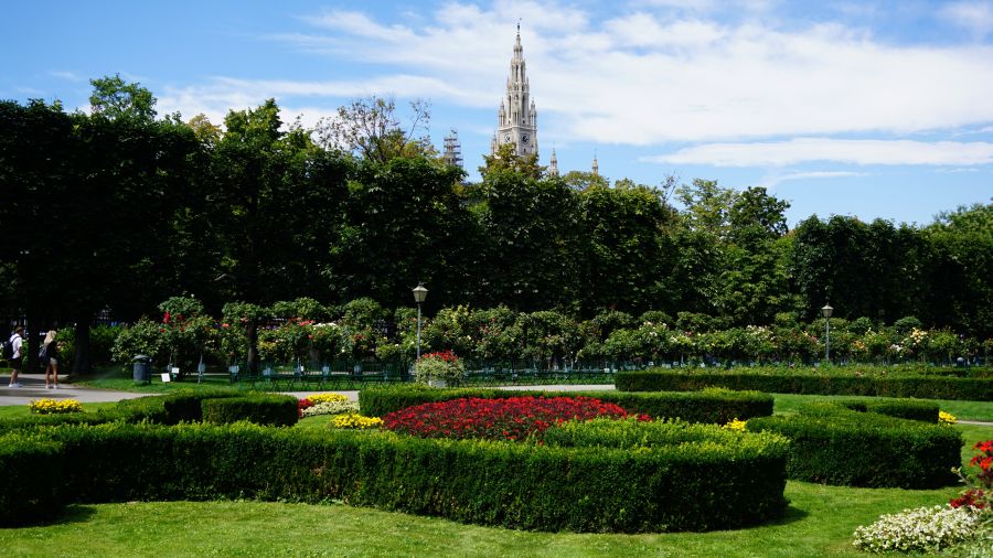 Blick auf gepflegte Gärten mit farbenfrohen Blumenbeeten und grünen Hecken im Vordergrund, im Hintergrund ist ein beeindruckender Kirchturm unter blauem Himmel sichtbar.