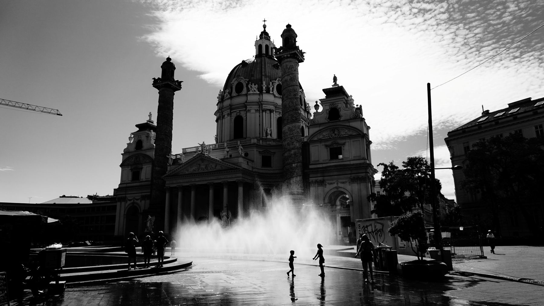 Schwarz-weiß Foto der Karlskirche in Wien. Im Vordergrund spielende Kinder vor einem Wasserspiel.