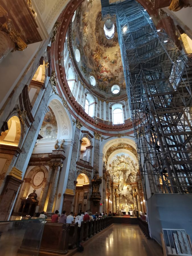 Blick in das Innere der Karlskirche in Wien, mit beeindruckender Kuppelmalerei und Baugerüst für Renovierungsarbeiten, während Menschen in den Bänken sitzen.