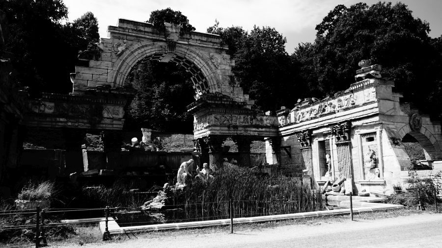 Eine Schwarz-Weißaufnahme einer monumentalen architektonischen Ruine mit einem großen Arkadenbogen, umgeben von Pflanzen und Bäumen.