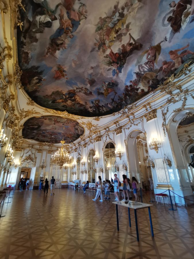 Innenansicht eines prächtigen Saals mit dekorativem Deckengemälde, Kronleuchtern und Gästen, die durch den Raum gehen.