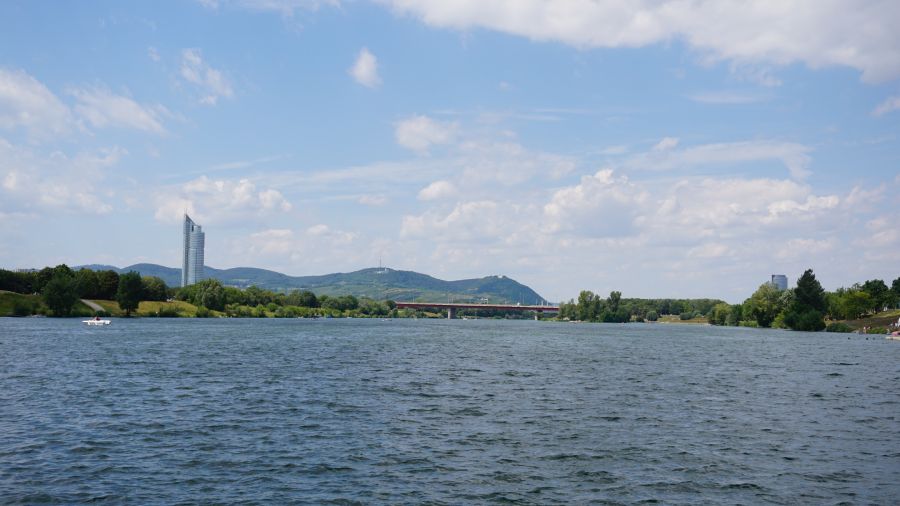 Ausblick auf die Donau mit einem klaren blauen Himmel und vereinzelten Wolken, im Hintergrund sind Bäume und ein Hochhaus zu sehen.