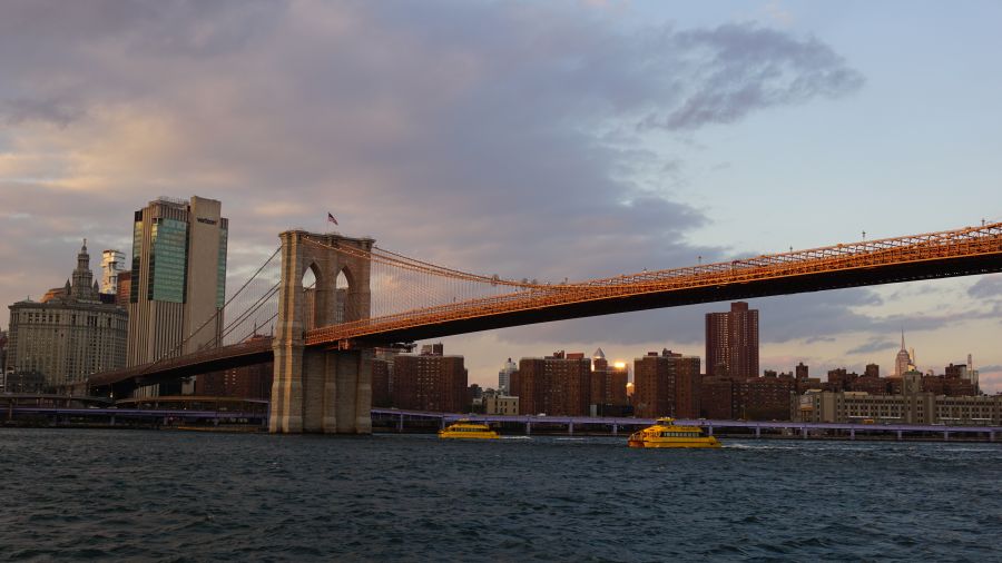 Aussicht auf die Brooklyn Bridge bei Sonnenuntergang, umgeben von der New Yorker Skyline und einem ruhigen Gewässer.