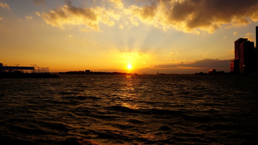 Sonnenuntergang über einer Wasserfläche mit strahlendem Licht und Wolken am Himmel, im Hintergrund schemenhafte Silhouetten von Gebäuden.