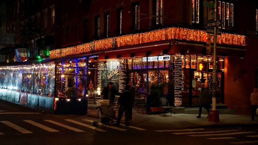 Beleuchtetes Restaurant mit festlicher Weihnachtsdekoration in einer Straßenszene, Menschen gehen vorbei.