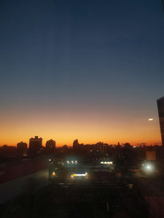 Blick aus dem Fenster auf die Skyline von New York City während des Sonnenaufgangs, mit orangefarbenem Himmel und beleuchteten Gebäuden im Vordergrund.