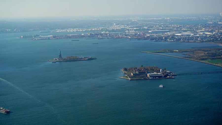 Luftaufnahme von Liberty Island mit der Freiheitsstatue und Ellis Island im Hintergrund, umgeben von Wasser und Booten.