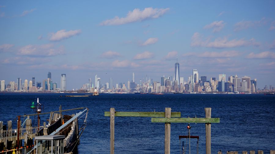 Blick auf die Skyline von New York City vom Wasser aus, mit dem One World Trade Center sichtbar in der Mitte und einem klaren blauen Himmel.
