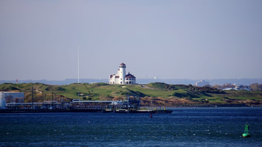 Blick auf einen Leuchtturm auf einer grünen Landzunge, umgeben von Wasser und einem klaren Himmel.