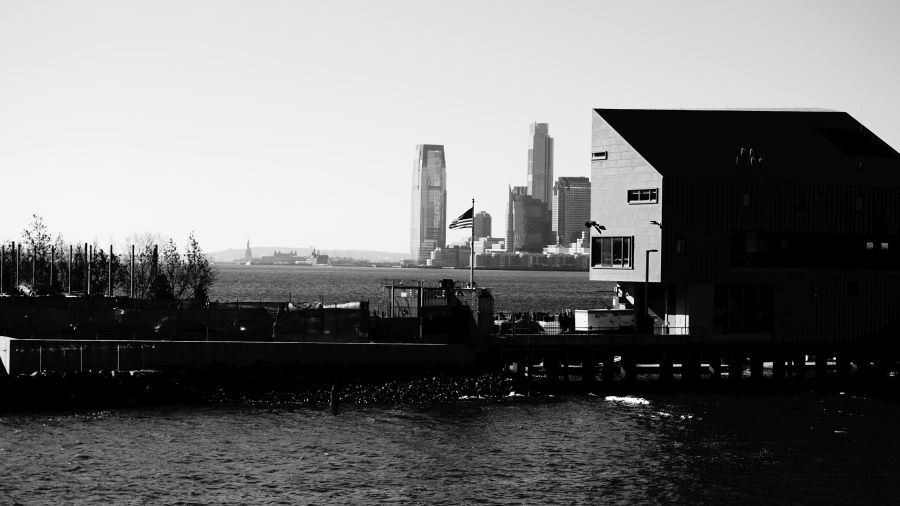 Blick auf den Hudson River mit moderner Architektur von Manhattan im Hintergrund, im Vordergrund ein Pier und eine Flagge wehend.