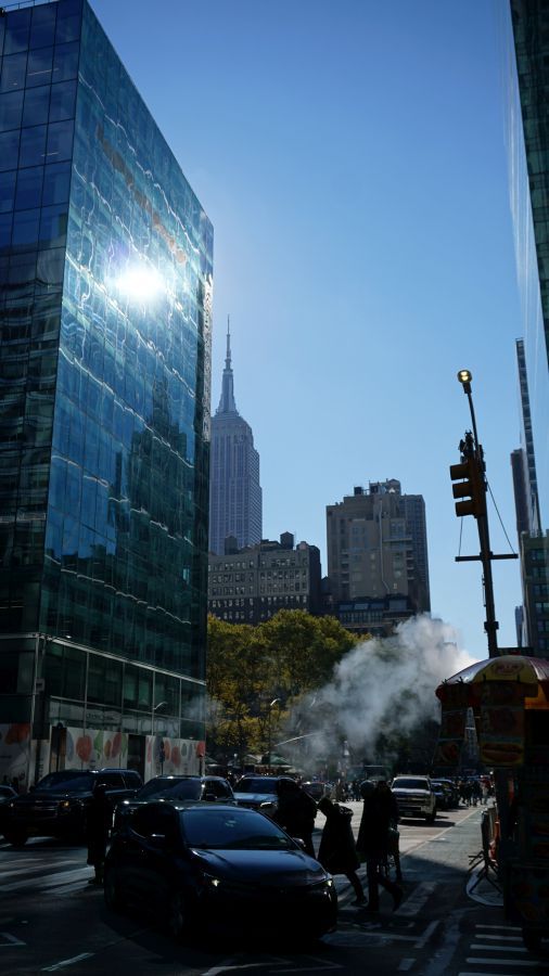 Blick auf die Straßen von New York mit dem Empire State Building im Hintergrund und modernen Glasgebäuden, bei klarem blauen Himmel.