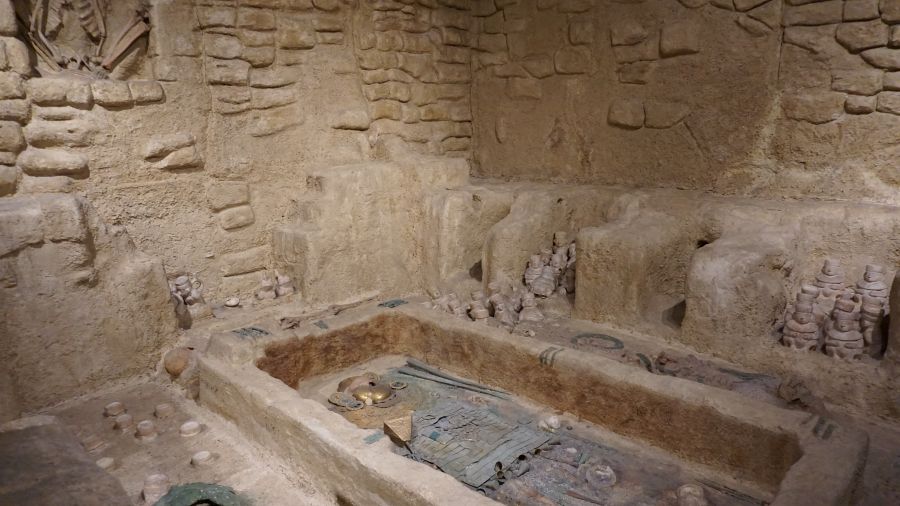 Einblick in eine archäologische Stätte mit einem Grabraum, umgeben von steinernen Wänden und verschiedenen Artefakten.