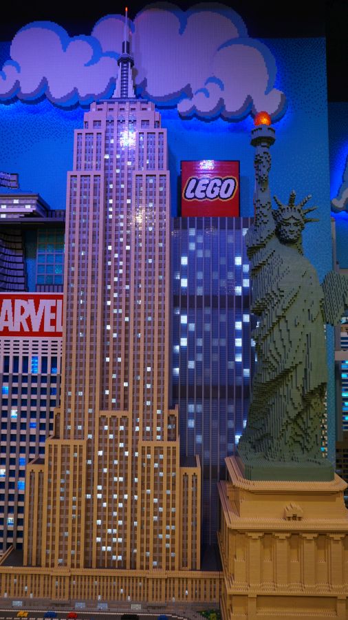 Eine LEGO-Darstellung des Empire State Buildings neben der Freiheitsstatue, umgeben von bunten LEGO-Elementen und Werbeschildern.