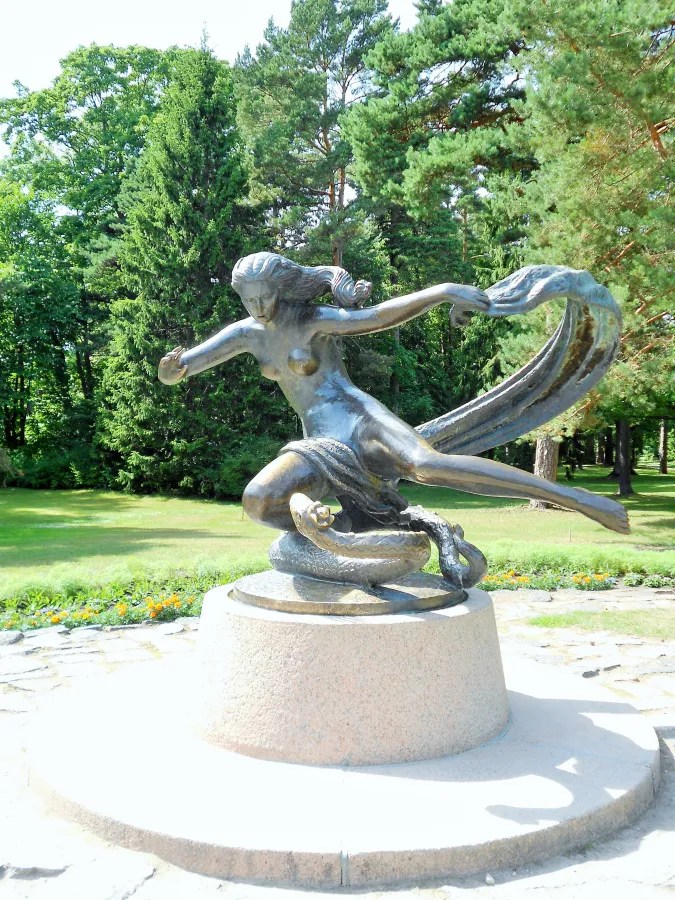 A bronze statue of a woman depicted in a flowing pose, holding a fabric while standing on a base, surrounded by greenery.