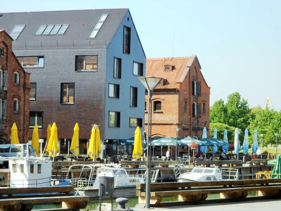 Hafenansicht mit modernen und historischen Gebäuden, gelben Sonnenschirmen und Booten im Wasser.