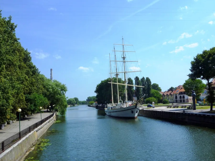 Ein Segelschiff auf einem ruhigen Kanal, umgeben von Bäumen und Uferpromenade, unter einem blauen Himmel.