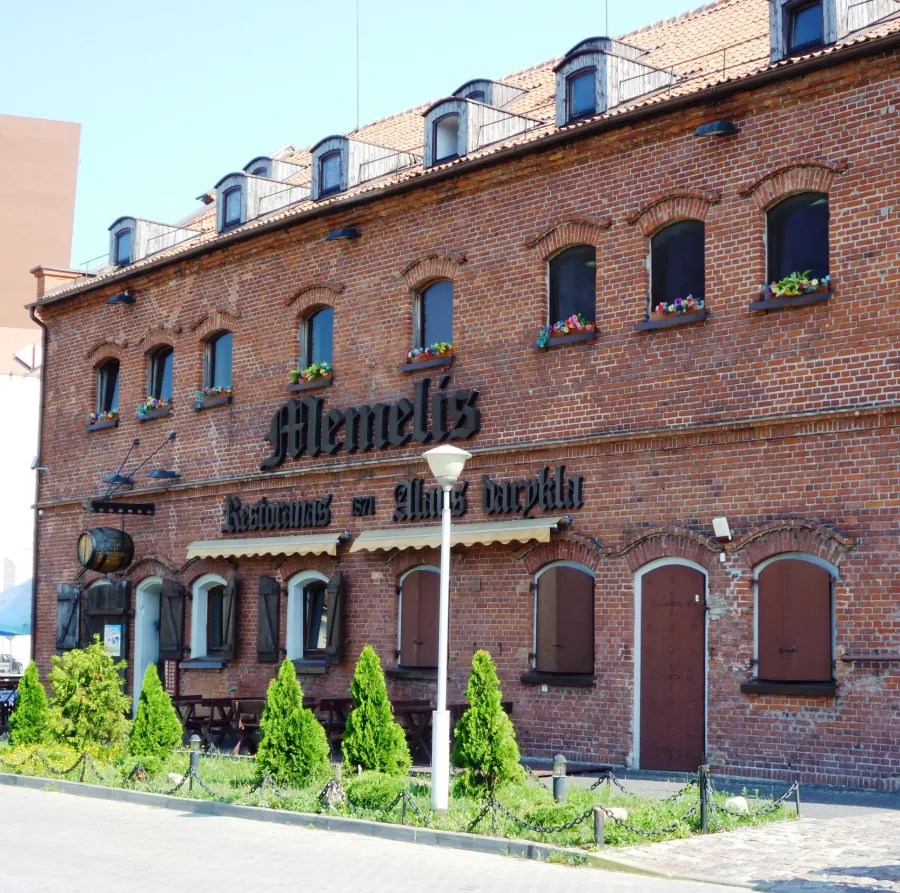 Exterior view of the Memelis restaurant in Klaipeda, showcasing its brick facade, windows decorated with flowers, and surrounding greenery.