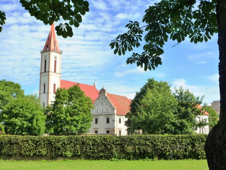 A picturesque view of a building with a tall steeple surrounded by lush green trees and a hedge, under a blue sky with scattered clouds.