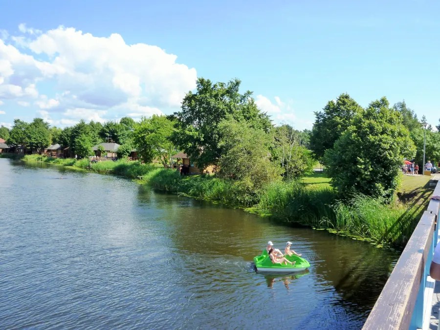 Two people kayaking in a green pedal boat on a peaceful river surrounded by lush trees and wooden cabins.