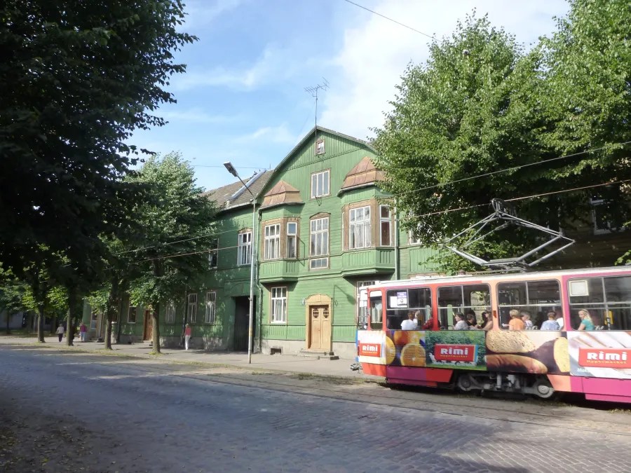 Eine grüne Holzgebäude mit mehreren Fenstern und einem Eingangsbereich, in der Nähe einer Straßenbahn, unter blauen Himmel und umgeben von Bäumen.