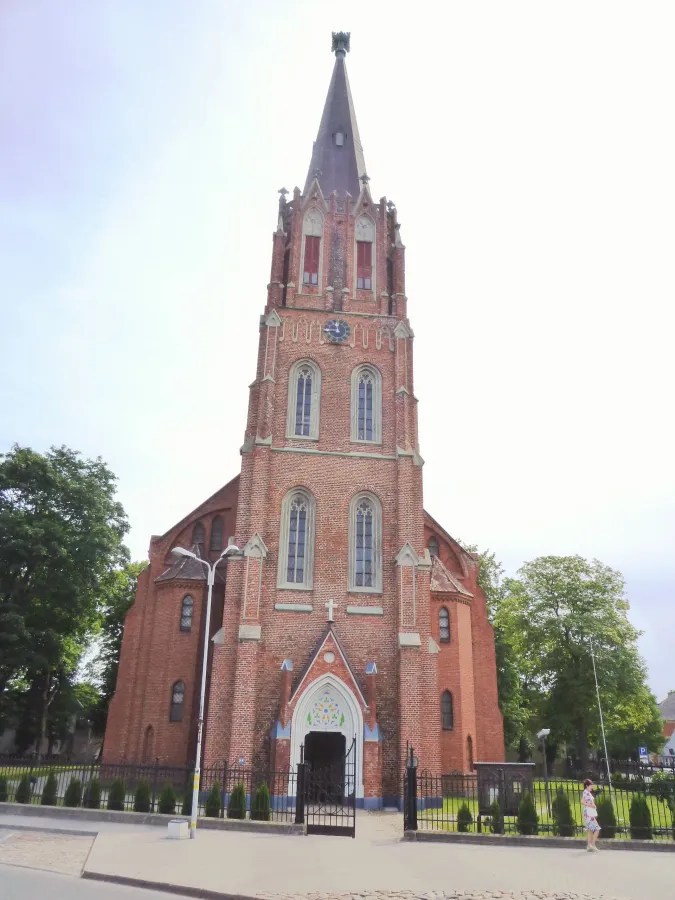Ein historisches Backsteingebäude mit einem hohen, spitzen Turm und bunter Fensterverzierung, umgeben von Bäumen und einem Zaun.