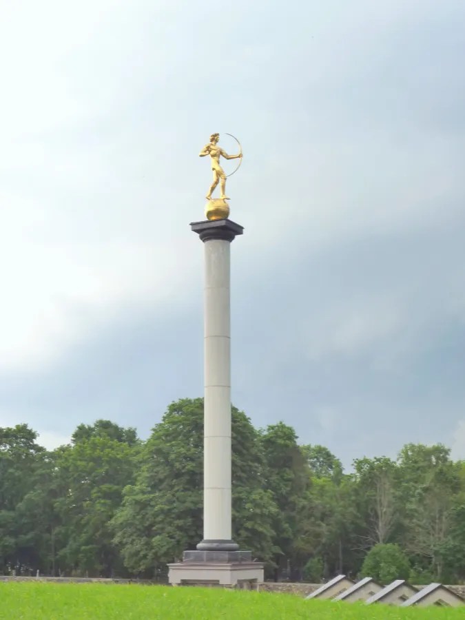 Goldene Statue auf einer hohen Säule im Freien, umgeben von Bäumen und Gras, unter einem bewölkten Himmel.