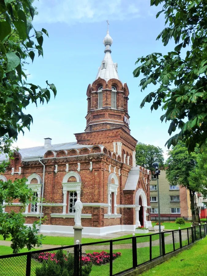 Historische Ziegelkirche mit einem hohen Turm und einer Statue im Vordergrund, umgeben von Bäumen und Blumenbeeten.