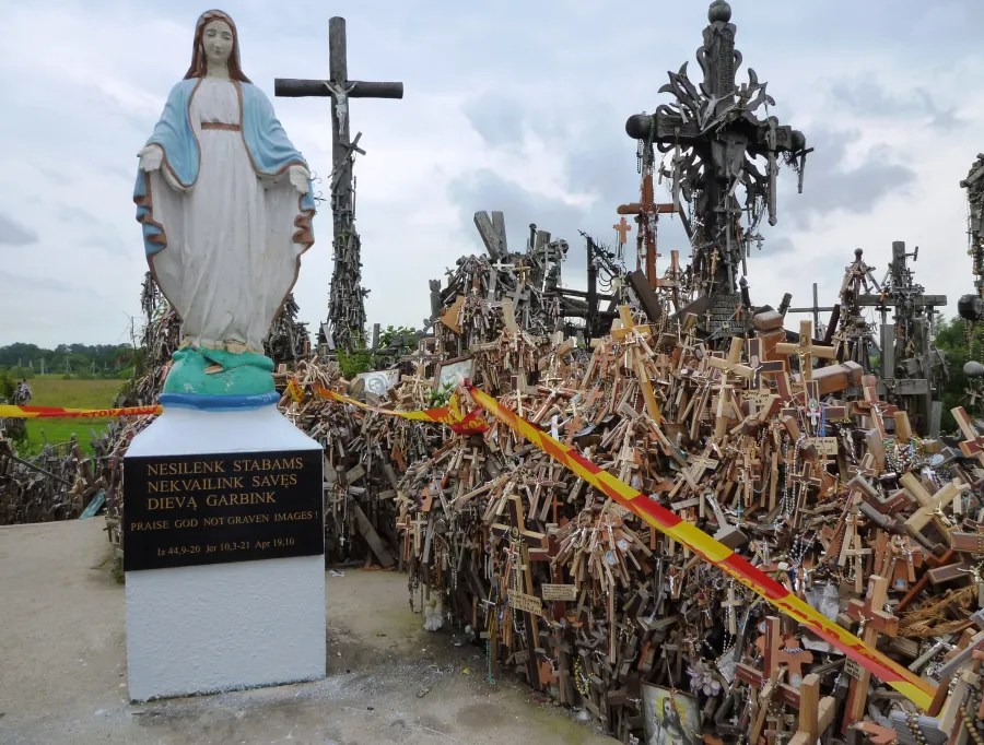 Statue der Jungfrau Maria vor dem Berg der Kreuze, umgeben von zahlreichen Holz- und Metallkreuzen, unter einem bewölkten Himmel.