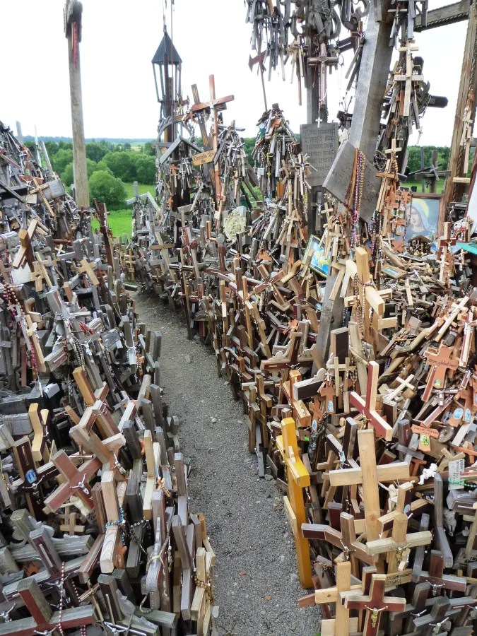 Eine enge Gasse zwischen vielen Holz- und Metallkreuzen, die auf einem Hügel aufgestellt sind. Im Hintergrund sind grüne Felder sichtbar.
