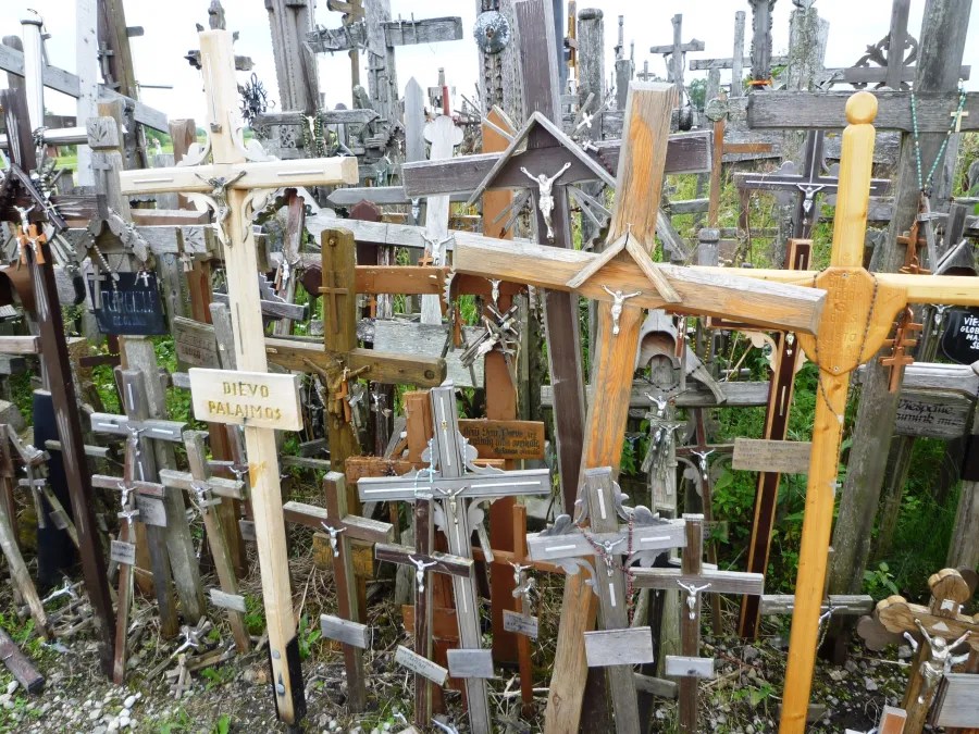 Ein überfüllter Bereich mit verschiedenen Holz- und Metallkreuzen, die in chaotischer Anordnung stehen, umgeben von Grün.