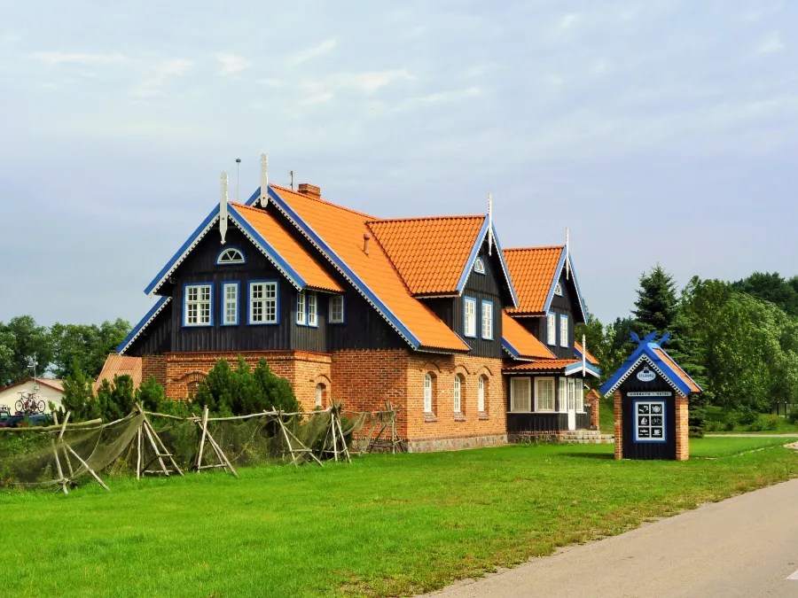 A traditional two-story house with a reddish-orange tiled roof, featuring dark wooden siding and decorative blue accents, set in a green landscape.