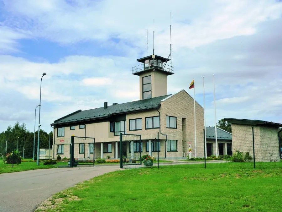 A modern airport building with a control tower, surrounded by greenery and a fenced area.