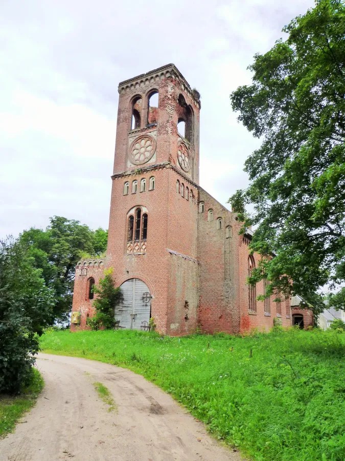 Eine verlassene Backsteinruine mit einem hohen Turm und großen Fenstern, umgeben von grüner Vegetation und einem gravelweg.