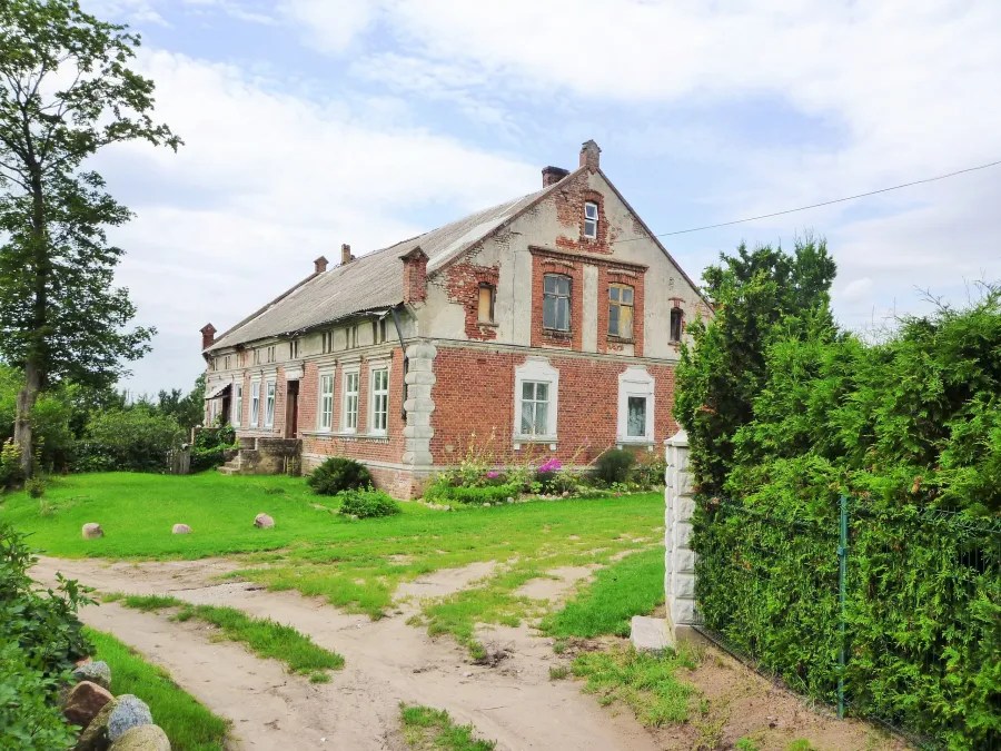 A weathered, historic building with a mix of brick and plaster exterior set in a green landscape with a gravel path and trees.