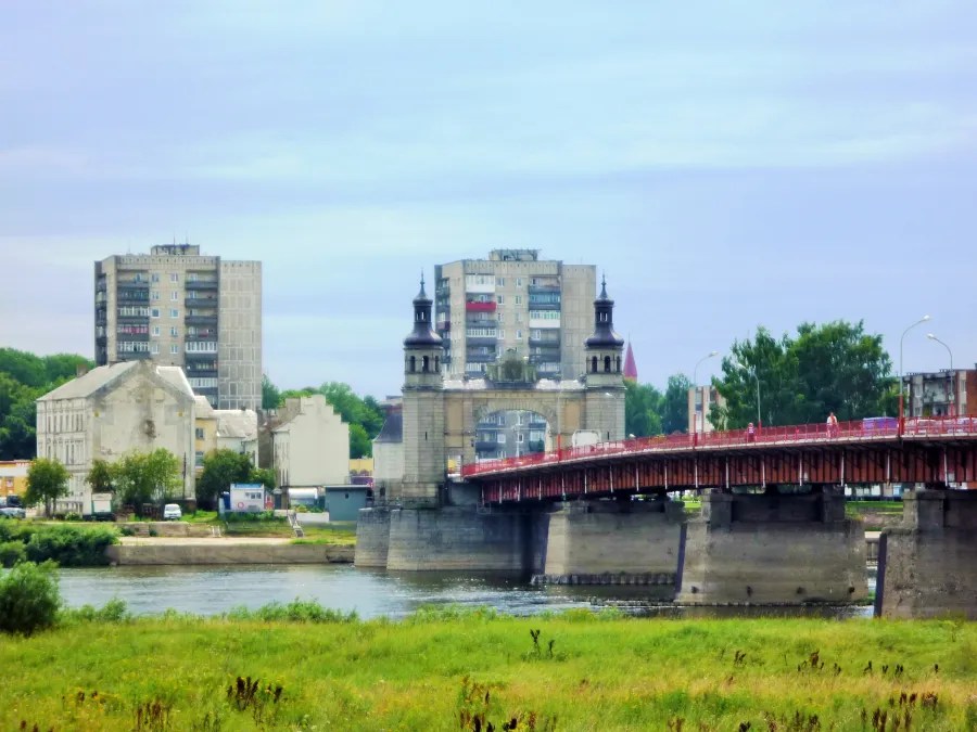 Blick auf eine Ziegelsteinbrücke mit Türmen, umgeben von modernen Wohngebäuden und grüner Landschaft.